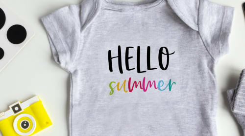 Flatlay image of a light fabric transfer design with black and rainbow font reading, 'hello summer,' applied to a heather gray onesie, with a yellow camera and other items surrounding it.