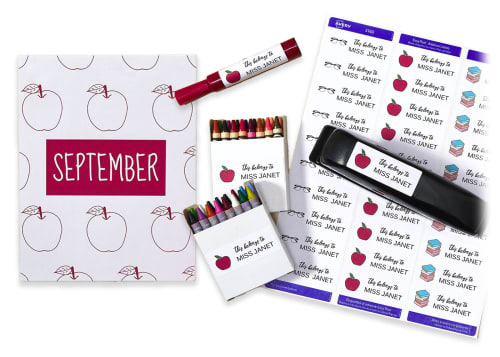 Flatlay of various school supplies, including a folder, crayon boxes, a marker, and a stapler, next to a sheet of printable labels, with multiple designs printed on the labels themselves.