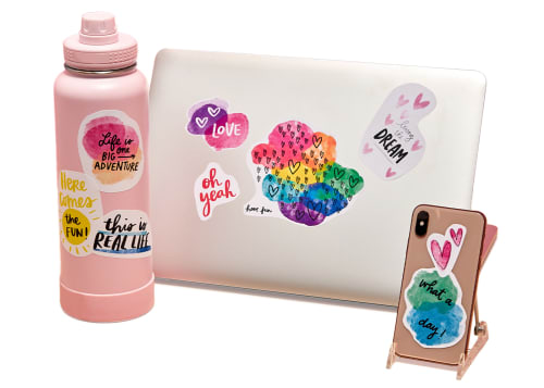 A laptop, a pink reusable water bottle, and a phone on a stand, each covered in various removable decals
