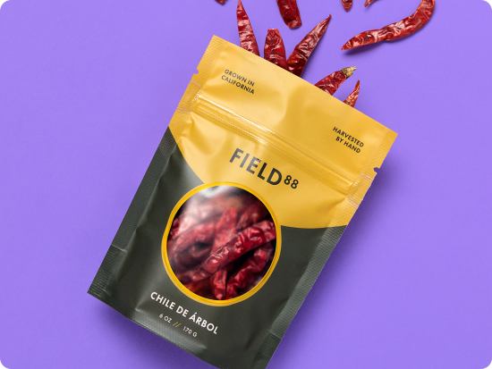 A custom pouch with a dark forest green and bright sunny yellow design, containing chiles from a business called Field88. The pouch is laying on a bright purple background, with dried chiles coming out of the back, strwen across the surface.