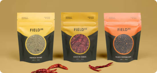 Three stand-up pouches of different seasonings sitting on a brown background with dried chiles around them. One contains thyme, another has dried chiles, and the last holds black peppercorn.