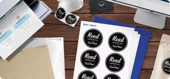 Flatlay of sheets of labels with a black and white design, next to a printer, sheets of blank labels, business cards, and a computer.