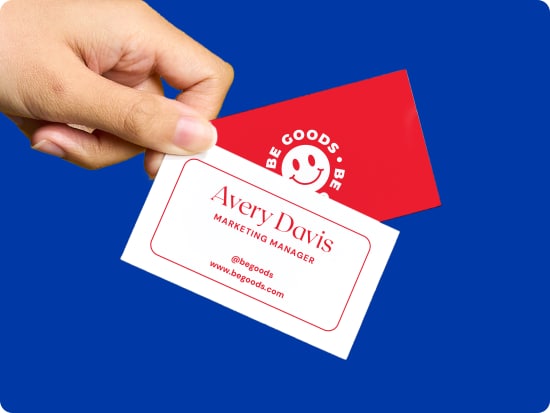 Two busness cards, one showing the front of the design with red text on a white background, and one showing the back of the design with a red background and a smiley-face logo that says 'Be Goods.' The cards are held and fanned out by their corners by a hand, against a deep blue background.