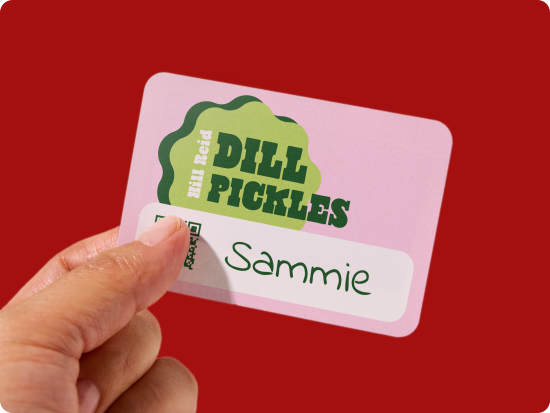 A light pink name tag with a cute pickle slice design being held up by a person's hand against a deep red background.