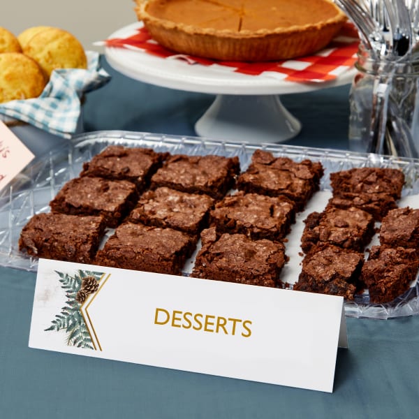 A tray of brownies are set on a potluck buffet table. In front of the brownies is a large Avery tent card 5305 printed with a evergreen foliage and pine cones. The card reads, "Desserts."
