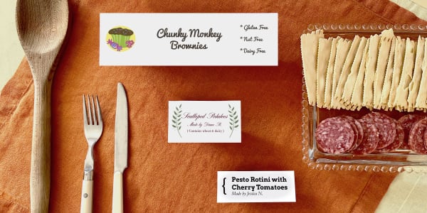 Three different sizes of food tent cards are shown on a table next to a wooden spoon, knife, fork and snacks for size reference. The three food tent cards shown are Avery 5303 which is 2-1/2 inches by 8 inches, Avery 5302 which is 2 inches by 3-1/2 inches, and Avery 16109 which is 1-7/16 inches by 3-3/4 inches.