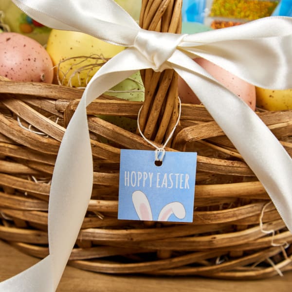 Close up of brown wicker Easter basket with elegant spotted Easter eggs and decorated with a white satin bow and "Hoppy Easter" Avery tag 22849. 