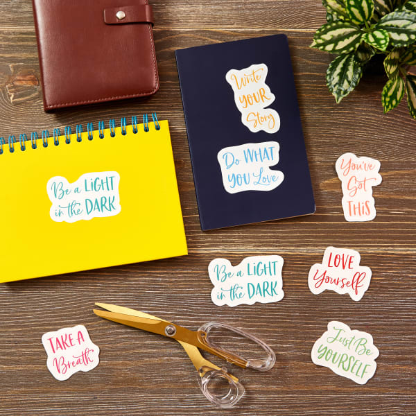 Motivational sticker templates for making your own decals. The decals are shown cut out and arranged on a wooden table near some scissors, a plant and journals. 