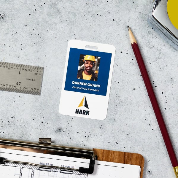 A photo ID name tag for work showing dark blue background with an employee photo and name as well as a company logo. The name tag is made of durable plastic with a hang hole to use with a clip or lanyard. 