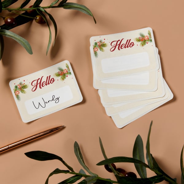 A classy Christmas party name tag idea featuring a subtle creamy, beige background with holiday greenery accents and cheerful red lettering that reads, "Hello." There is white space to handwrite individual names.