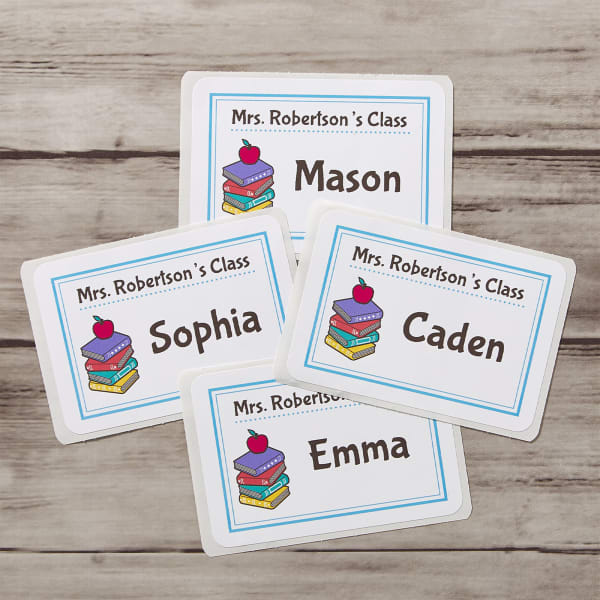 A classic book-themed name tag idea for school. The design features a spot to add the teacher's class name and the student name in a casual and friendly font in a handwritten style. The text is accented with a cute graphic depicting a stack of books with an apple on top. 