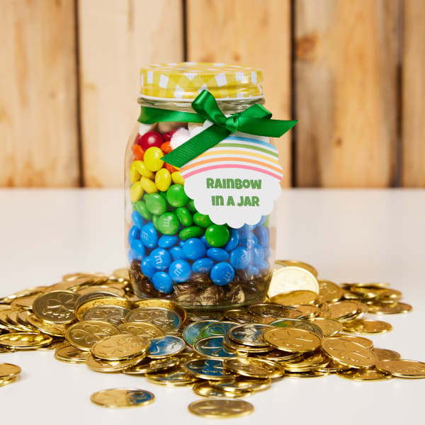An upcycled jam jar with yellow gingham-print lid is filled with rainbow candies and decorated with a green ribbon and rainbow tag. Avery 80511 tag reads, "Rainbow in a Jar."