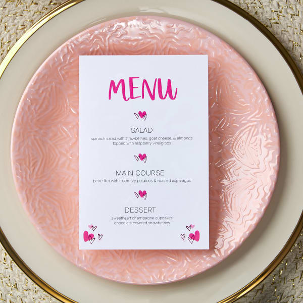 A free Avery template for a party menu is shown on Avery postcard 5389. The template features bright pink text and bright pink hearts that contrast nicely with an elegant light pink, cream, and gold table setting.