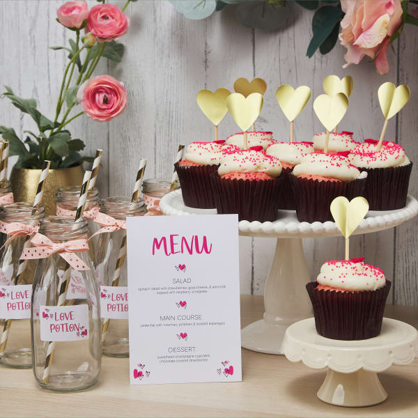 A demonstration of how to set up a buffet table at a DIY Galentine's Day party. The set up includes a DIY menu card, "love potion" drink bottles, and cupcake toppers made with blank gold Avery heart-shaped labels.