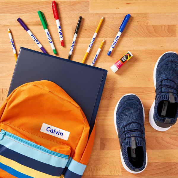 A kids backpack and shoes on a wooden floor. The backpack is open with a binder, markers, and a glue stick spilling out. the backpack, markers and inside the shoes are labeled with Avery no-iron fabric labels 40720.