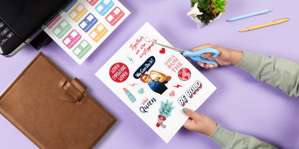 A top down image of someone cutting out decorative DIY planner stickers with scissors. The table top is lavender and there is a printer with more DIY planner stickers coming out as well as a leather planner and some décor accents. 