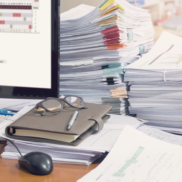 A messy home office desk with stacks of paperwork cluttering the desktop and crowding the computer.