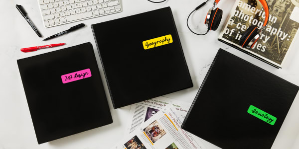 A grouping of economy binders are shown on a college desktop. The binders are personalized with various stickers. 