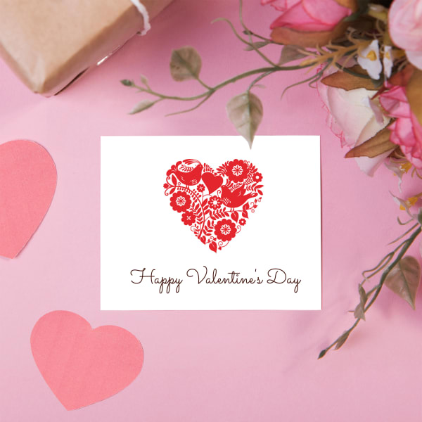 Avery 3378 half-fold greeting card is shown on a pink table with a bouquet of flowers. The card features a Valentine's Day Template with a red botanical-themed heart shape. 