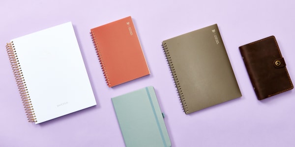 Bullet journals laid out on a plain lavender background for size comparison. Sizes A5 (5.8 x 8.3 inches), A6 (4.1 x 5.8 inches), and A4 (8.3 x 11.7 inches) are shown. 