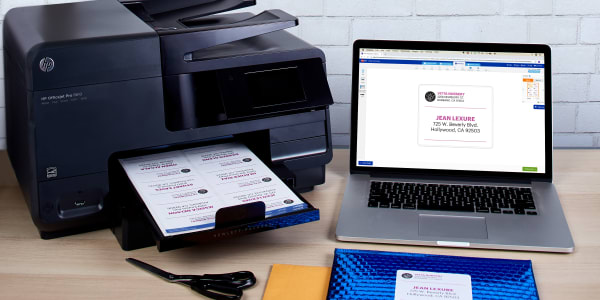 A picture of a plain wooden desktop with a laptop, printer, scissors and shipping envelopes. The laptop is open and shows the editing screen in Avery Design and Print Online for customizing a shipping label. There are freshly-printed Avery labels with Sure Feed technology in the printer tray and one is applied to a metallic blue bubble mailer envelope. 