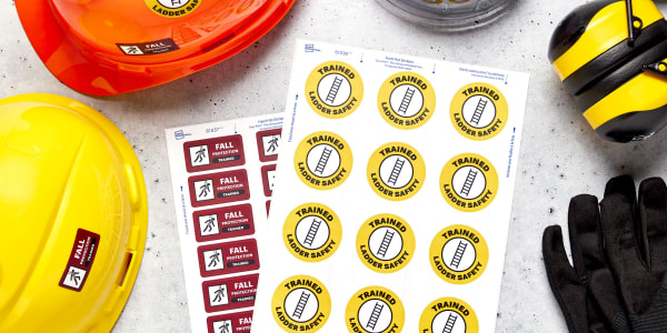 Two sheets of Avery hard hat stickers are shown on a table surrounded by construction PPE, including hard hats, gloves, and safety ear muffs. The labels shown are Avery 61536, which are round hard hat stickers, and Avery 61537, which are rectangle hard hat stickers.