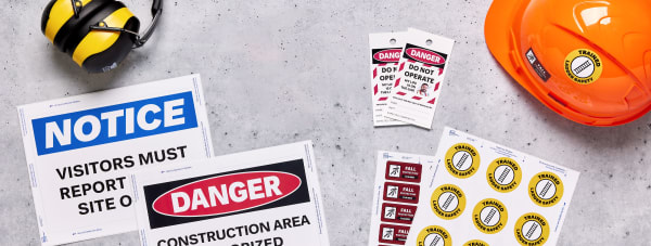 Flatlay of construction safety materials and equipment, including a hard hat, sign labels, safety tags, and more, on a concrete surface