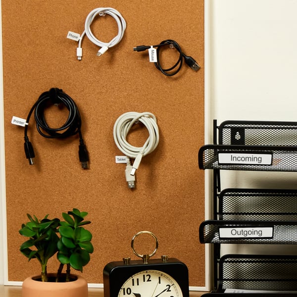 A cork board with device cables neatly hung up on push pins. Each cord is labeled with Avery barbell labels that indicate what device they go with.