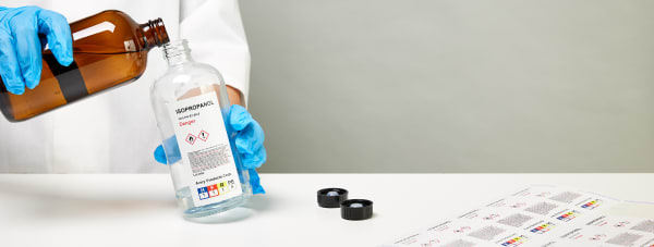 Laboratory personnel pouring fluid from a brown glass bottle into a clear bottle labeled wit ha GHS-compliant label. A sheet of these labels sits off the the side on a white table.