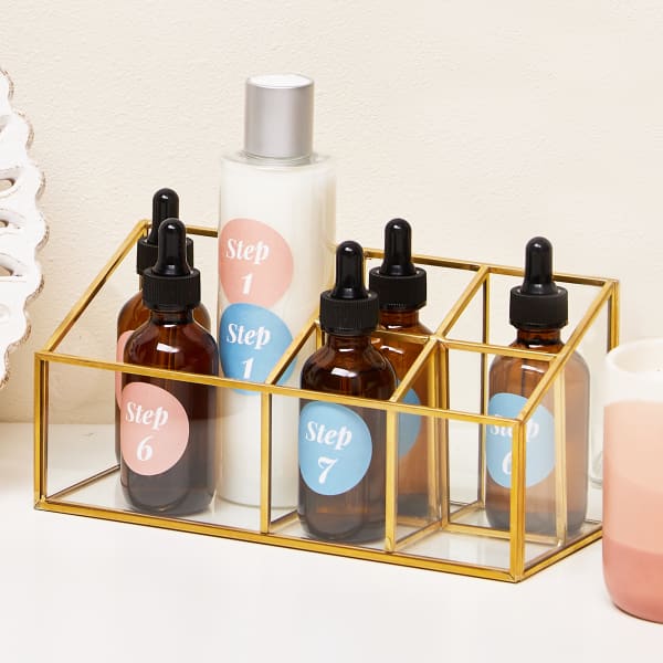 Counter-top skincare organization for day and night routines. Cool apothecary dropper bottles and a larger face lotion bottle are color-coded with pink and blue labels for morning and evening use. All the bottles are neatly stored in a modern clear organizer with graphic gold trim.