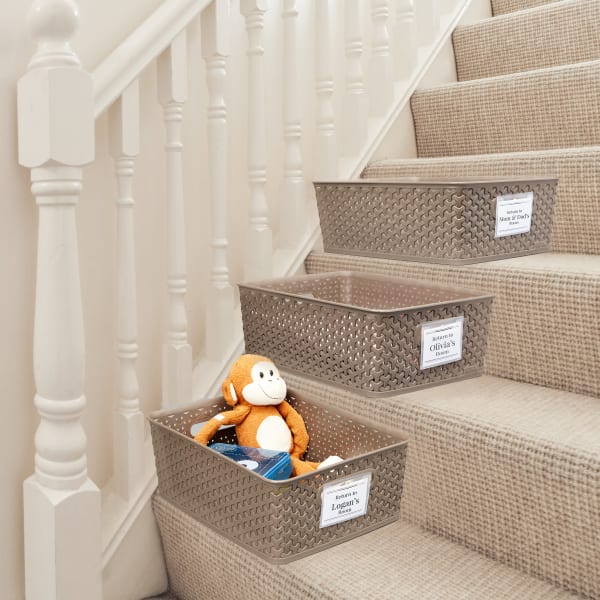 Personal clutter baskets staged on a staircase to help a family declutter their home. The baskets have Avery name badges 74651 clipped on them with different family members' names. 