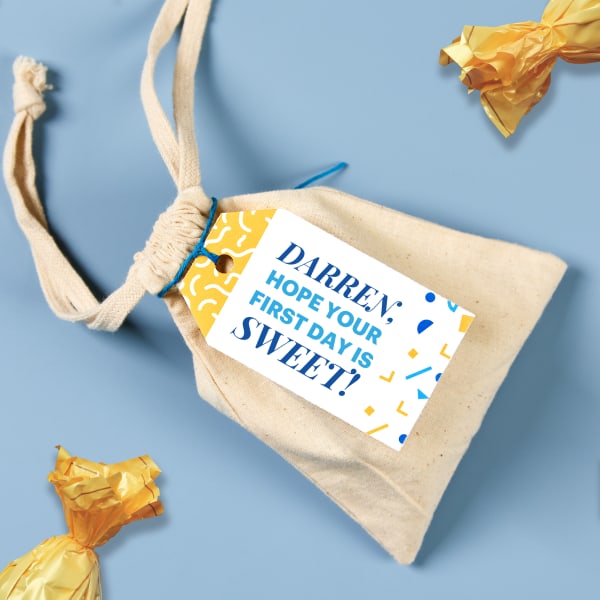 A small canvas bag and candy on a blue table. The canvas bag is personalized with Avery tag 22802 and reads, "Darren; hope your first day is sweet!"