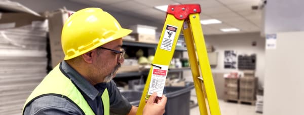 Person in safety gear reading a ladder inspection tag that is attached to a yellow and red ladder