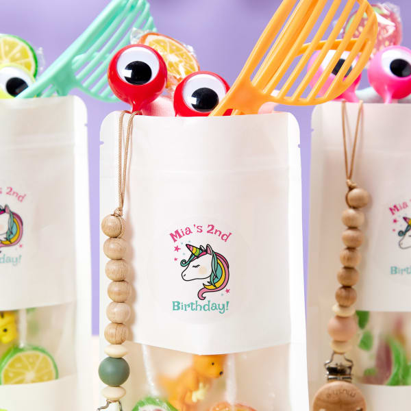 A close-up of DIY party favor bags for a birthday. The plain bags have personalized stickers applied, which read "Mia's 2nd Birthday" with a rainbow unicorn icon. The stickers are made with Avery 22830 labels and a free Avery template.