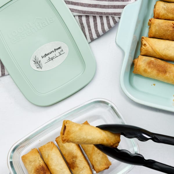 This is an example of a party planning label for packing up leftovers. This image shows a tray of eggrolls being transferred to a glass container with a plastic lid. The lid shows Avery label 4223 printed with a leaf design and the words "Leftovers from:" next to a space to fill in by hand.