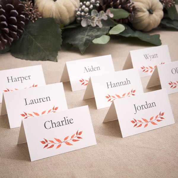 Eight place cards for Thanksgiving set out on a table. They feature a simple orange leaf design and are personalized with individual guest names. 