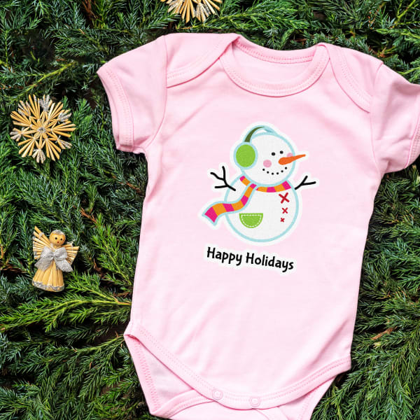 Image showing a pink baby onesie with a holiday template of a snowman and text that reads “Happy Holidays” printed on a glitter fabric transfer.