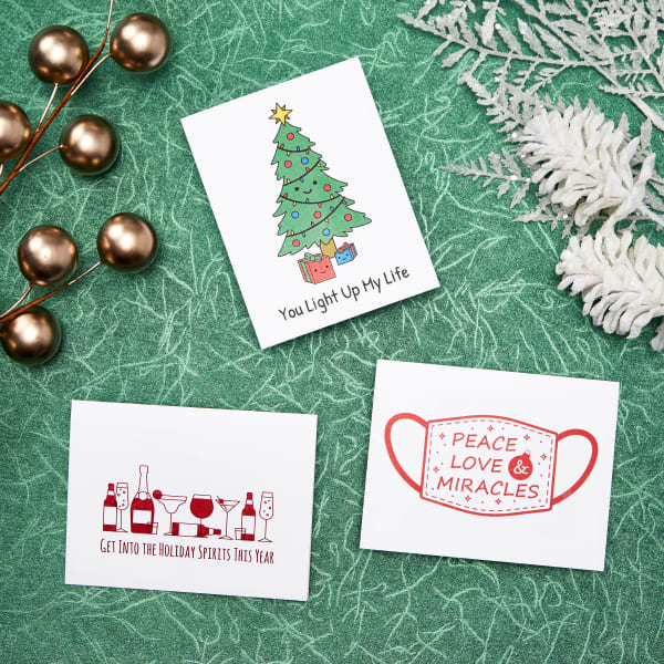 Three holiday cards made with Avery note card 8315. One has a Christmas tree and reads, " You Light Up My Life." Another shows wine and cocktails and reads, "Get into the holiday spirits this year." The last card has a drawing of a face mask with the words, "Peace, Love & Miracles."