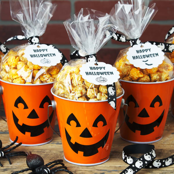 A group shot of three jack o' lantern buckets filled with caramel corn . Each bucket is topped with a black and white ghost ribbon and black and white tag that reads "Happy Halloween."