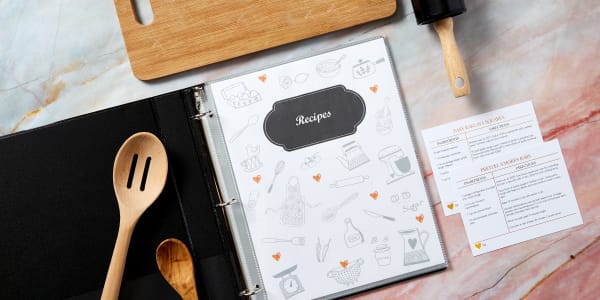 Contemporary modern marble counter top with a DIY recipe book and recipe cards artistically laid out with cooking utensils and a cutting board. The recipe book features an Avery binder and sheet protectors.