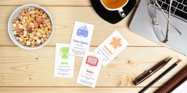 Avery printable business cards printed with Avery templates for team building icebreaker cards with colorful graphics. The cards are laid out on a rustic wood desk and surrounded by a laptop, snacks and desk supplies.