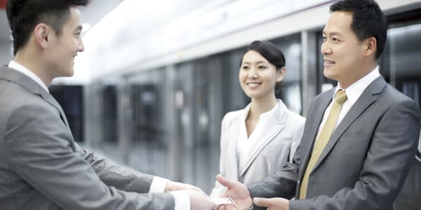 Three business professionals in formal attire are engaged in a friendly exchange. Two men are shaking hands and exchanging business cards, while a woman stands nearby, smiling and observing. The setting appears to be a modern office or corporate environment, emphasizing a formal and professional interaction.