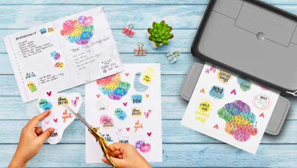 a persons hands demonstrating how to make stickers in custom sizes using avery sticker project paper with bright fun planner stickers and an open bullet journal planner and avery sticker paper coming out of a desktop printer shown on a pretty blue wooden background