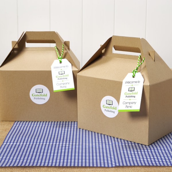 Company picnic idea for a boxed picnic lunch with branding. Two cardboard lunch boxes are shown on blue checkered paper. Each box has a custom logo sticker on the front and a welcome tag with the company's logo on it. 