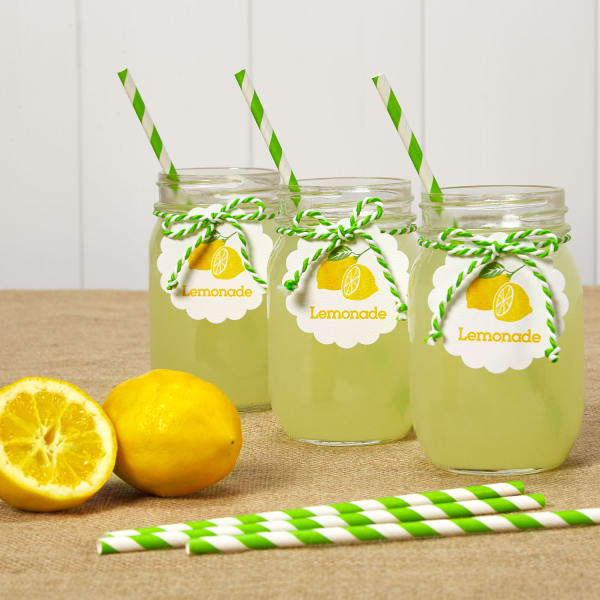 Three Mason jars are filled with lemonade and decorated with lemonade tags and green striped paper straws. The Mason jars are arranged on a table with fresh lemons and more straws. 