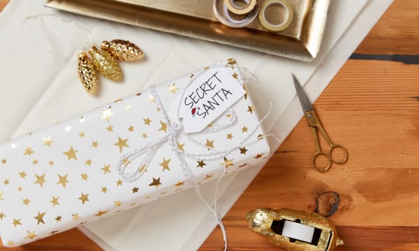 wooden table surface with holiday gift wrapping material including wrapping paper scissors tape ribbon and wrapped gift with secret santa avery gift tag