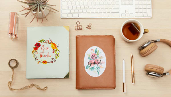 Leatherbound gratitude journals sitting on a wood desk with a warm cup of tea, a pair of headphones and various office supplies