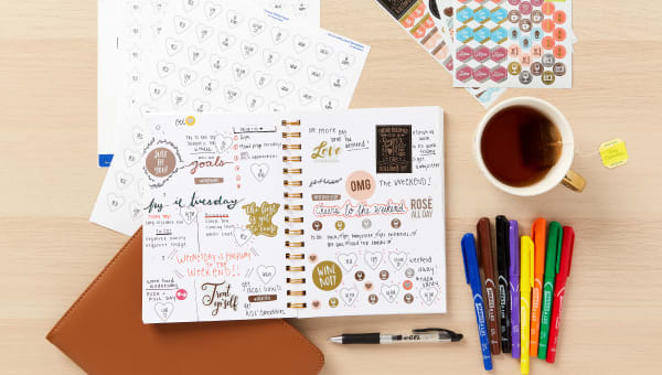 A gratitude journal sitting on a wood table along with custom-printed stickers, permanent markers and a cup of tea