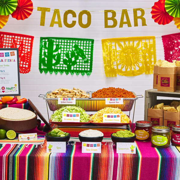 A taco bar is set up on a colorful sarape-inspired table cloth. The food is labeled with Avery place cards and tent cards personalized with cactus, floral, and papel picado designs. 