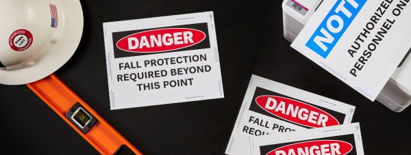 Flatlay of three preprinted danger signs next to a white hard hat and a white printer, printing out a notice sign.